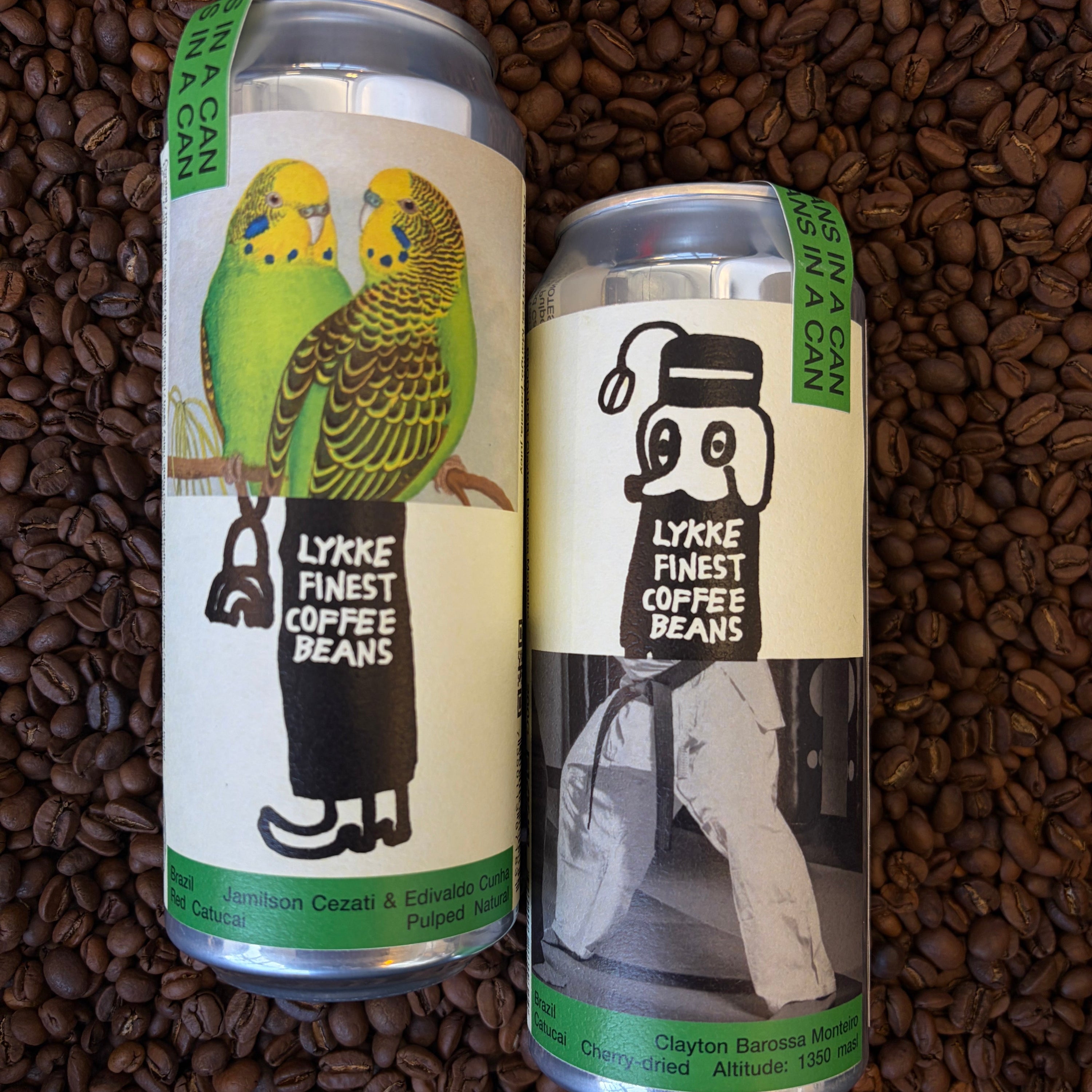 Two aluminum cans of Lykke Finest Coffee Beans placed on a bed of dark roasted coffee beans. The left can features a white label with green accents and an illustration of two green budgerigars, while the right can has a black-and-white label with a cartoon character wearing a hat and scarf.